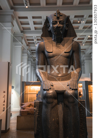 A dark stone statue of an Egyptian pharaoh seated on a throne, displayed in a London museum with high ceilings, columns, and display cases in the background. A dark stone statue of an Egyptian pharaoh seated on a throne, displayed in a London museum with high ceilings, columns, and display cases in the background. 130807933