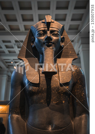 A detailed statue of an Egyptian pharaoh with a headdress and false beard, displayed indoors with a coffered ceiling and museum surroundings in London. 130807938
