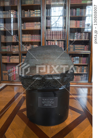 The Rosetta Stone rests on a black pedestal with an informational plaque, surrounded by bookshelves and wooden floors, likely in the British Museum. 130808007