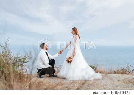 Groom kneeling in front of bride by the sea Groom kneeling in front of bride by the sea 130810425