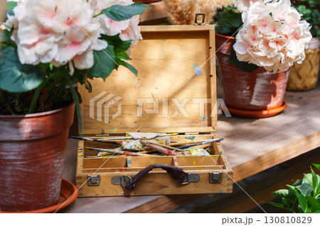 Old wooden paint box with brushes surrounded by blooming flowers in pots on sunny wooden table 130810829