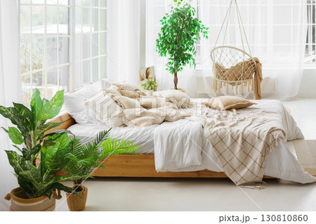 Comfortable bed with lots of soft decorative beige pillows in light sunny bedroom with large window 130810860