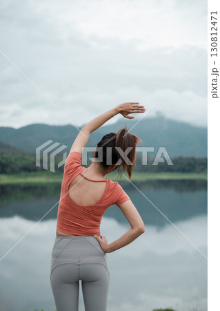 A woman in activewear stretches by a tranquil lake with mountains in the background, showcasing outdoor fitness and nature. 130812471
