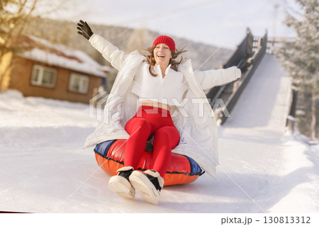 A young woman with a bright red beanie and matching leggings is joyfully tubing down a snowy slope. Dressed in a cozy white coat, she throws her arms wide open, embracing the thrill of winter sports. 130813312