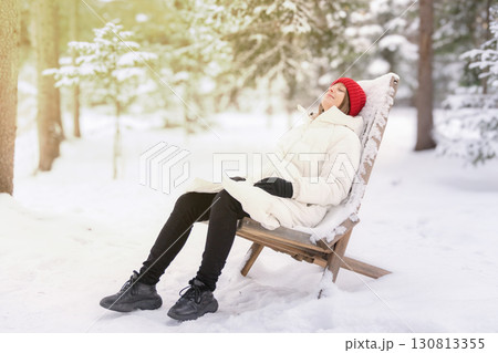A woman in a white down jacket sits reclining on a wooden armchair in a snowy forest. Relaxing in nature after the Christmas holidays 130813355