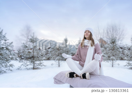 A woman in a white cap and scarf meditates peacefully in winter in nature against the background of trees on a winter Christmas morning. 130813361