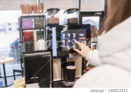 A woman in a white jacket buys a coffee drink or tea from a coffee machine at a gas station or store, selecting the desired drink on the screen with her finger.Close-up view from behind. 130813372
