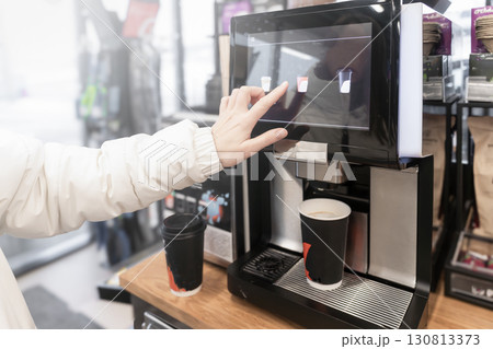 A woman pours and buys cappuccino coffee on her own at a self-service machine at a gas station or in a store. A close-up hand selects a drink with its finger on the touchscreen of a coffee machine. A woman pours and buys cappuccino coffee on her own at a self-service machine at a gas station or in a store. A close-up hand selects a drink with its finger on the touchscreen of a coffee machine. 130813373