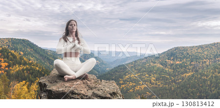 A woman in white leggings sits on a mountain in autumn in a yoga lotus position. Meditation practice on a mountain cliff during autumn surrounded by vibrant colors and serene landscapes A woman in white leggings sits on a mountain in autumn in a yoga lotus position. Meditation practice on a mountain cliff during autumn surrounded by vibrant colors and serene landscapes 130813412