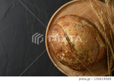 Top view of a freshly baked, crusty artisanal sourdough loaf, presented on a rustic wooden plate. A serrated knife rests beside the bread, with a bunch of golden wheat ears arranged elegantly on the 130814089