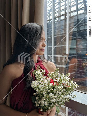 Young beautiful woman in a red evening dress with a bouquet thoughtfully looks out the window 130817171