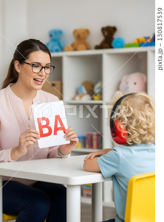 Autistic child boy with noise cancelling headphone in speech therapy practicing phonics with therapist holding letter card for communication skills and autism awareness 130817539