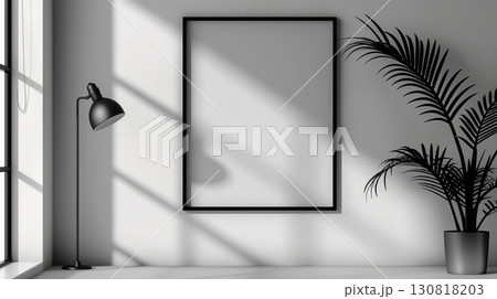 Black frame with a blank white canvas on a white wall with a floor lamp and a green plant in shadowed light 130818203