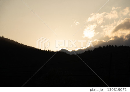 Golden sunlight peeks over mountain peaks at dusk, illuminating clouds and silhouetting trees in the valley 130819071