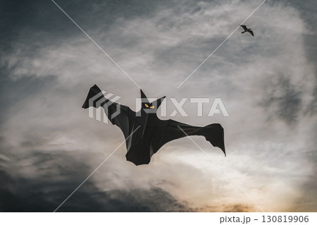 Black bat-shaped kite with glowing yellow eyes flying against dramatic evening sky, creating eerie and mysterious atmosphere. Halloween concept 130819906