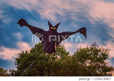 Dark bat-shaped kite with glowing yellow eyes flying against dramatic evening sky over trees, creating a spooky Halloween atmosphere 130819907