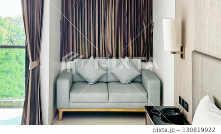 Interior of modern cozy hotel room with gray sofa, elegant dark curtains, simple bedside lamp, and serene window view revealing lush greenery, capturing feel of minimalist design and tranquil comfort 130819922
