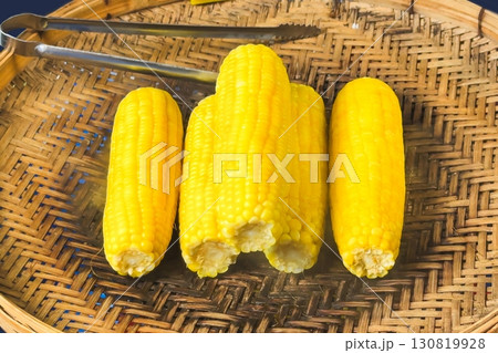 Boiled ears of corn rest on a woven bamboo tray, their bright yellow kernels offering a healthy, quick snack, embodying the simplicity and appeal of traditional street food 130819928