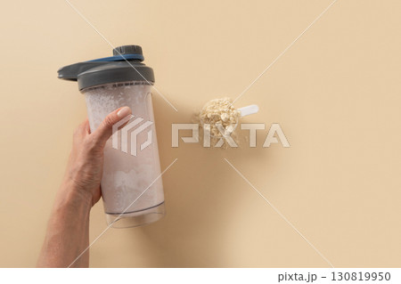 Whey protein shaker in hand and powder in scoop on beige background. Close up. 130819950