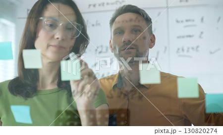 Professional woman with glasses marking glass board, arranging sticky notes while male colleague reviews strategic planning within modern, well lit workspace. Business people concept Professional woman with glasses marking glass board, arranging sticky notes while male colleague reviews strategic planning within modern, well lit workspace. Business people concept 130820274