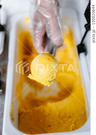 Hand scooping vibrant yellow mango gelato in a container, close-up food preparation 130820644