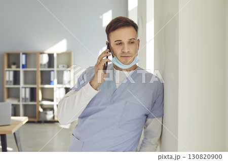 Male doctor is leaning on wall at clinic and calling someone on his mobile phone 130820900