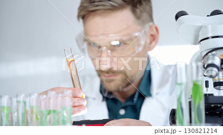 Man researcher with safety glasses is examining lab tube with a orange fluid in a modern laboratory, close-up. Science, microbiology and medicine 130821214