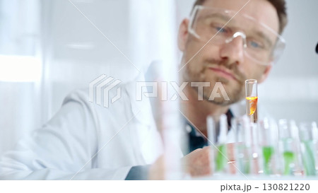 Man researcher with safety glasses is examining lab tube with a orange fluid in a modern laboratory, close-up. Science, microbiology and medicine 130821220