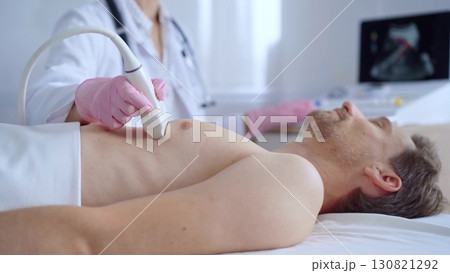 Doctor woman with pink medical gloves performing an ultrasound scan of male patient breast lying down in clinic. Cardiology diagnosis and medicine service concept Doctor woman with pink medical gloves performing an ultrasound scan of male patient breast lying down in clinic. Cardiology diagnosis and medicine service concept 130821292