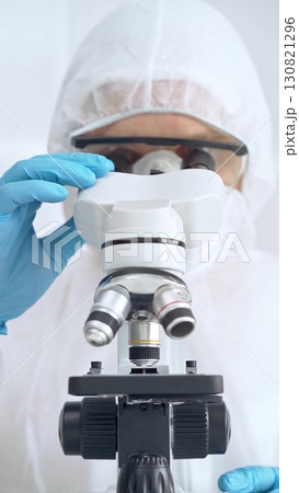 Scientist in ppe analyzing samples with microscope. Close-up of a focused researcher in a lab coat and protective gear in a laboratory setting Scientist in ppe analyzing samples with microscope. Close-up of a focused researcher in a lab coat and protective gear in a laboratory setting 130821296
