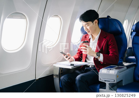 Asian businessman or entrepreneur in a formal suit on an airplane sitting in business class using a phone, computer laptop Asian businessman or entrepreneur in a formal suit on an airplane sitting in business class using a phone, computer laptop 130825249