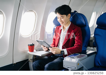 Asian businessman or entrepreneur in a formal suit on an airplane sitting in business class using a phone, computer laptop 130825250