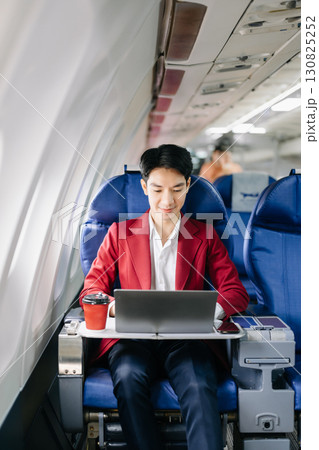 Asian businessman or entrepreneur in a formal suit on an airplane sitting in business class using a phone, computer laptop Asian businessman or entrepreneur in a formal suit on an airplane sitting in business class using a phone, computer laptop 130825252