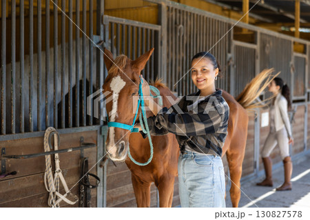Asian female worker takes horse out of stall, holds animal by bridle and leads pet for walk 130827578