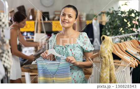 Near rack, asian woman buy summer clothes, choose some items. Female client looking for something in background 130828343