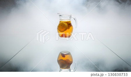 Transparent Glass Pitcher with Citrus Infused Drink Levitating Above Reflective Surface in White Backdrop 130830733