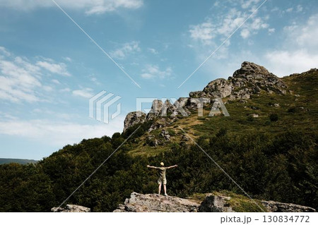A young woman raises her arms on a rock at Stara Planina national park, Serbia, with dramatic rocky slopes and lush green trees around, joy of mountain travel moment A young woman raises her arms on a rock at Stara Planina national park, Serbia, with dramatic rocky slopes and lush green trees around, joy of mountain travel moment 130834772