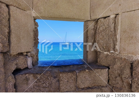Sea view from ancient wall, travel and vacation concept. Turquoise sea with sailboat, summer holiday, stone window to the ocean sea.  Rhodes Island Greece, Fortress in the old town. Sea view from ancient wall, travel and vacation concept. Turquoise sea with sailboat, summer holiday, stone window to the ocean sea.  Rhodes Island Greece, Fortress in the old town. 130836136