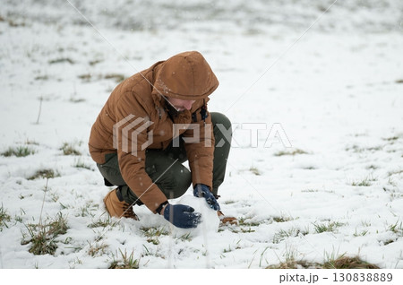 A person crouches in the snow, carefully shaping a small snowball. 130838889