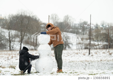 Father and son build a snowman together in a snowy landscape. A heartwarming winter scene. Father and son build a snowman together in a snowy landscape. A heartwarming winter scene. 130838891