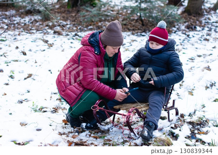 A woman and child use a phone together in a snowy forest, near a small wooden sled. 130839344