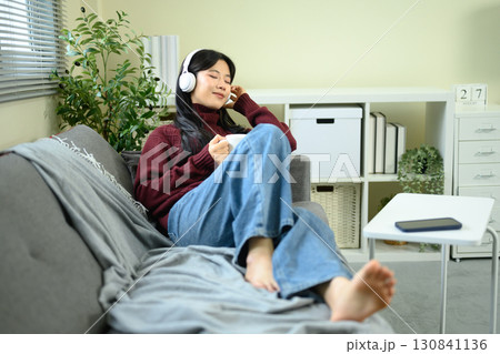 Young woman relaxing on the sofa, enjoying music with headphones and holding a cup of coffee 130841136