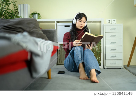 Young Asian woman sitting on the floor at home, wearing headphones and reading a book 130841149