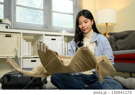 Young woman folding a beige sweater while packing her suitcase, preparing for an upcoming trip Young woman folding a beige sweater while packing her suitcase, preparing for an upcoming trip 130841178