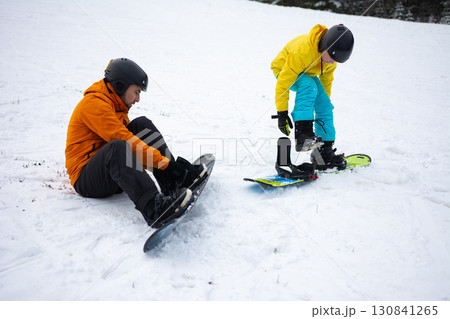 Two snowboarders prepare their equipment on a snowy slope surrounded by mountains during a sunny winter afternoon 130841265