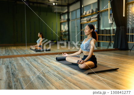 Young woman meditating in lotus position on yoga mat inside modern studio 130841277