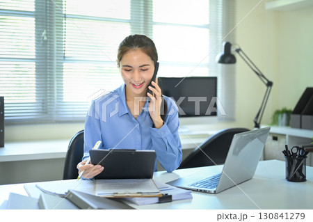 Smiling businesswoman talking on phone while reviewing documents in modern office 130841279