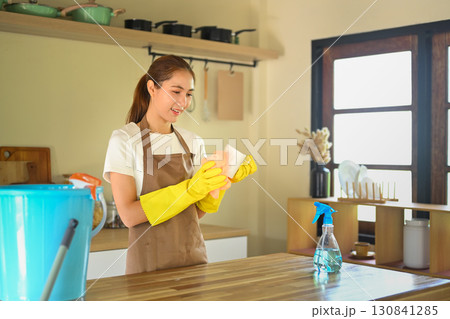 Young woman in apron and gloves wiping a cup with a cloth at a wooden kitchen counter Young woman in apron and gloves wiping a cup with a cloth at a wooden kitchen counter 130841285