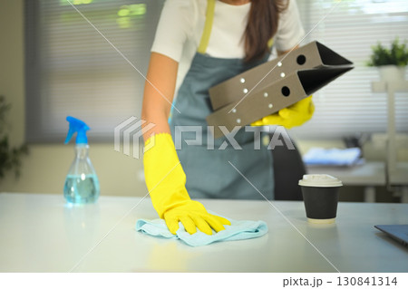 Female worker wearing apron and gloves wiping table with spray bottle and cloth in modern office Female worker wearing apron and gloves wiping table with spray bottle and cloth in modern office 130841314