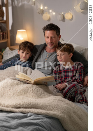 Father reads bedtime story to two children in cozy pajamas under warm blanket, creating peaceful and loving nighttime scene 130842838